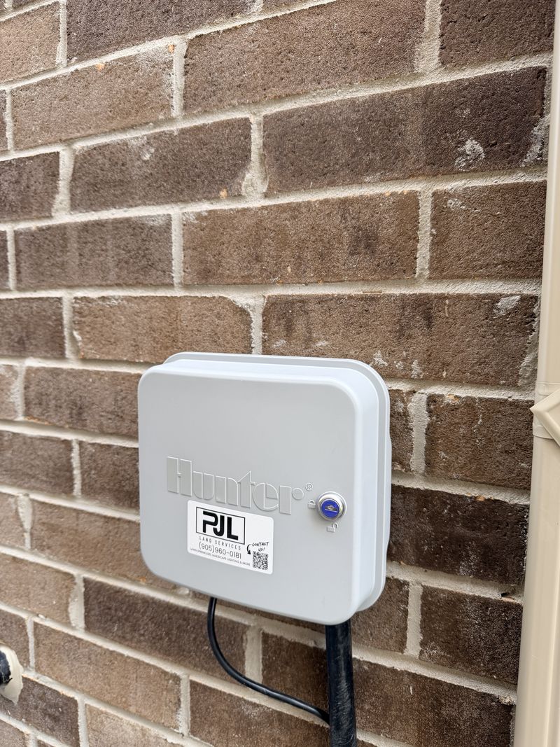 A Hunter sprinkler controller in a locked outdoor box on a brick wall, with a PJL sticker